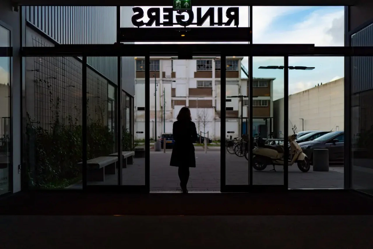 Person walking through a modern commercial building entrance with large glass doors and floor-to-ceiling windows.