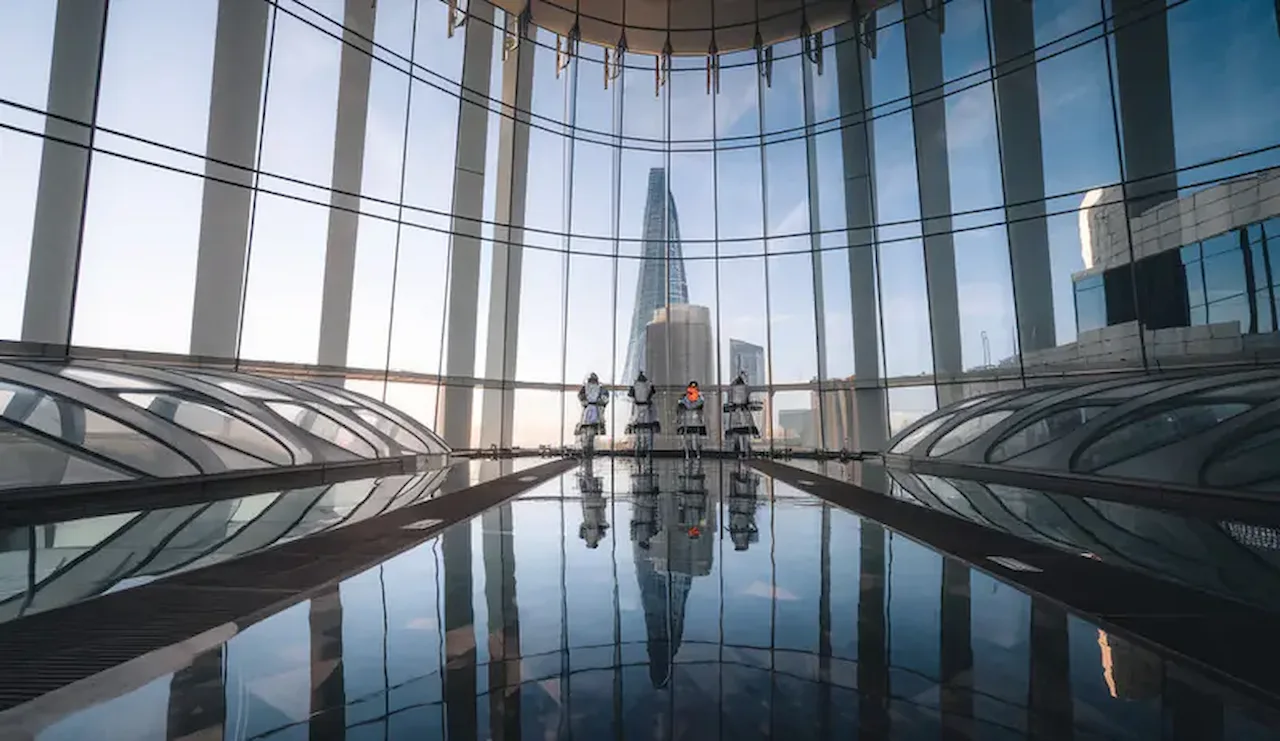 Spacious glass atrium with floor-to-ceiling windows, reflective flooring, and panoramic views of a modern city skyline.